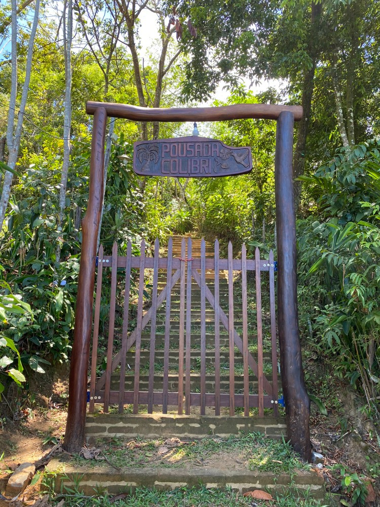 Portão de entrada da pousada entre árvores e vegetação.
